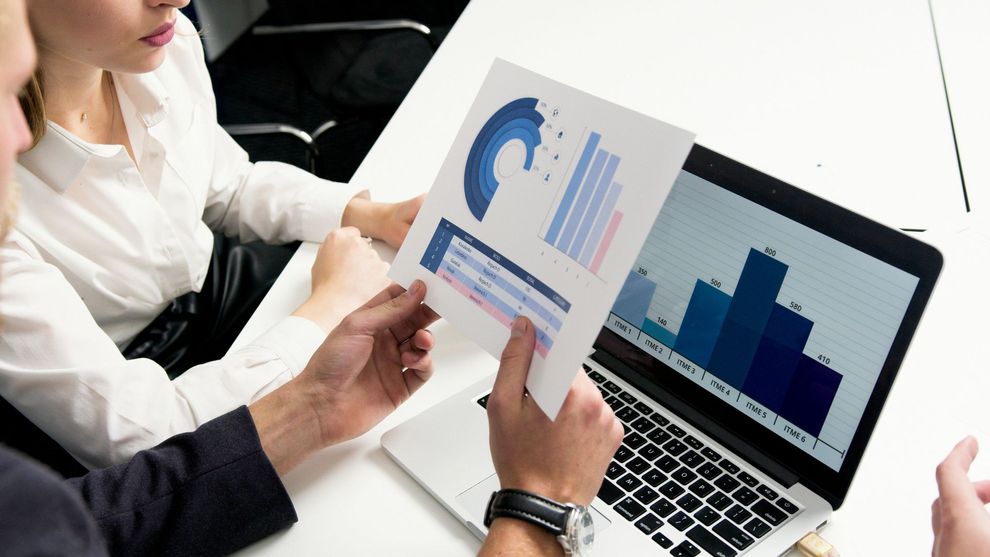 Two people analyze charts and graphs on paper and a laptop screen, possibly in a business meeting.