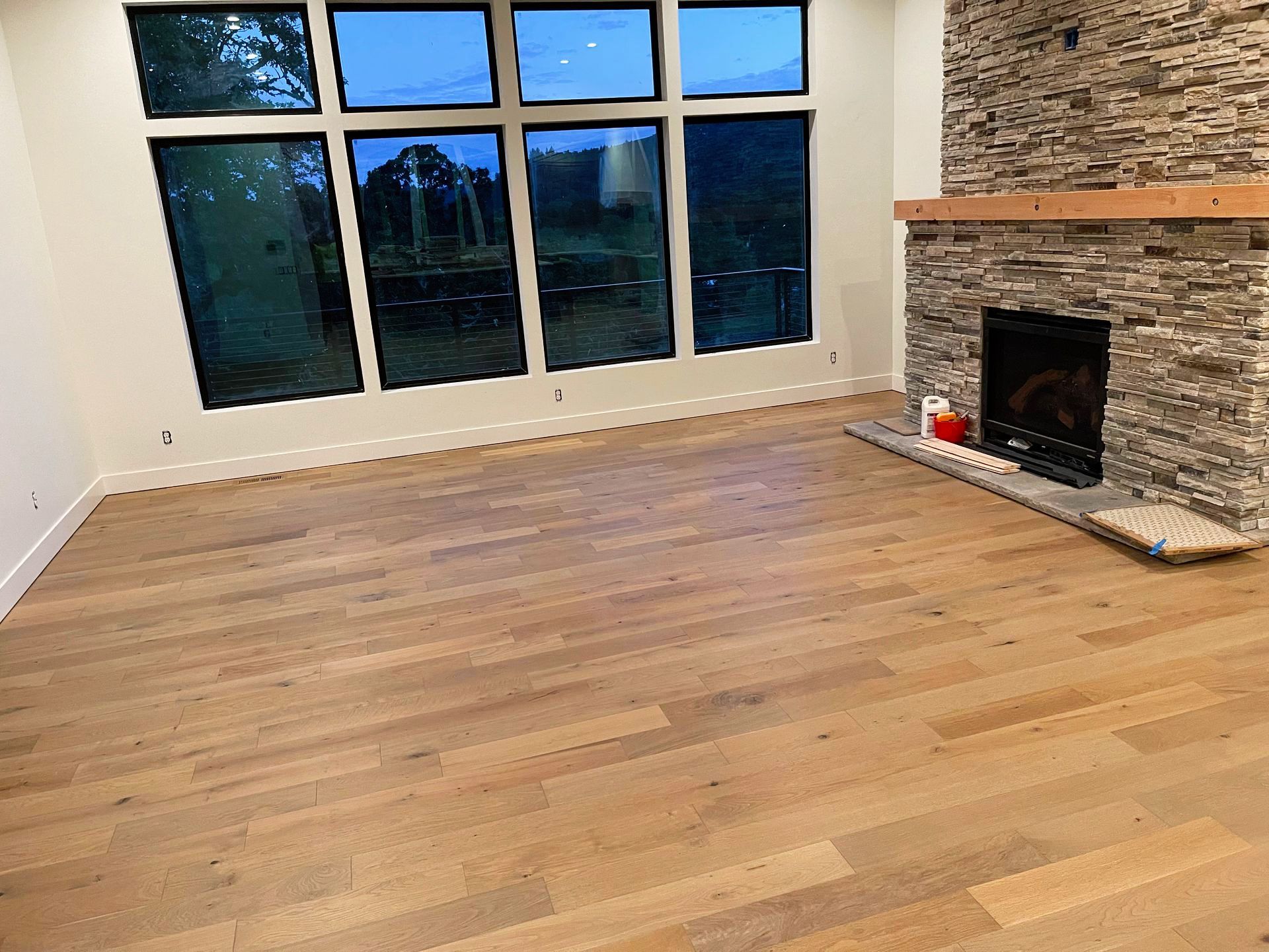 Empty room with hardwood floors, large windows, and a stone fireplace.