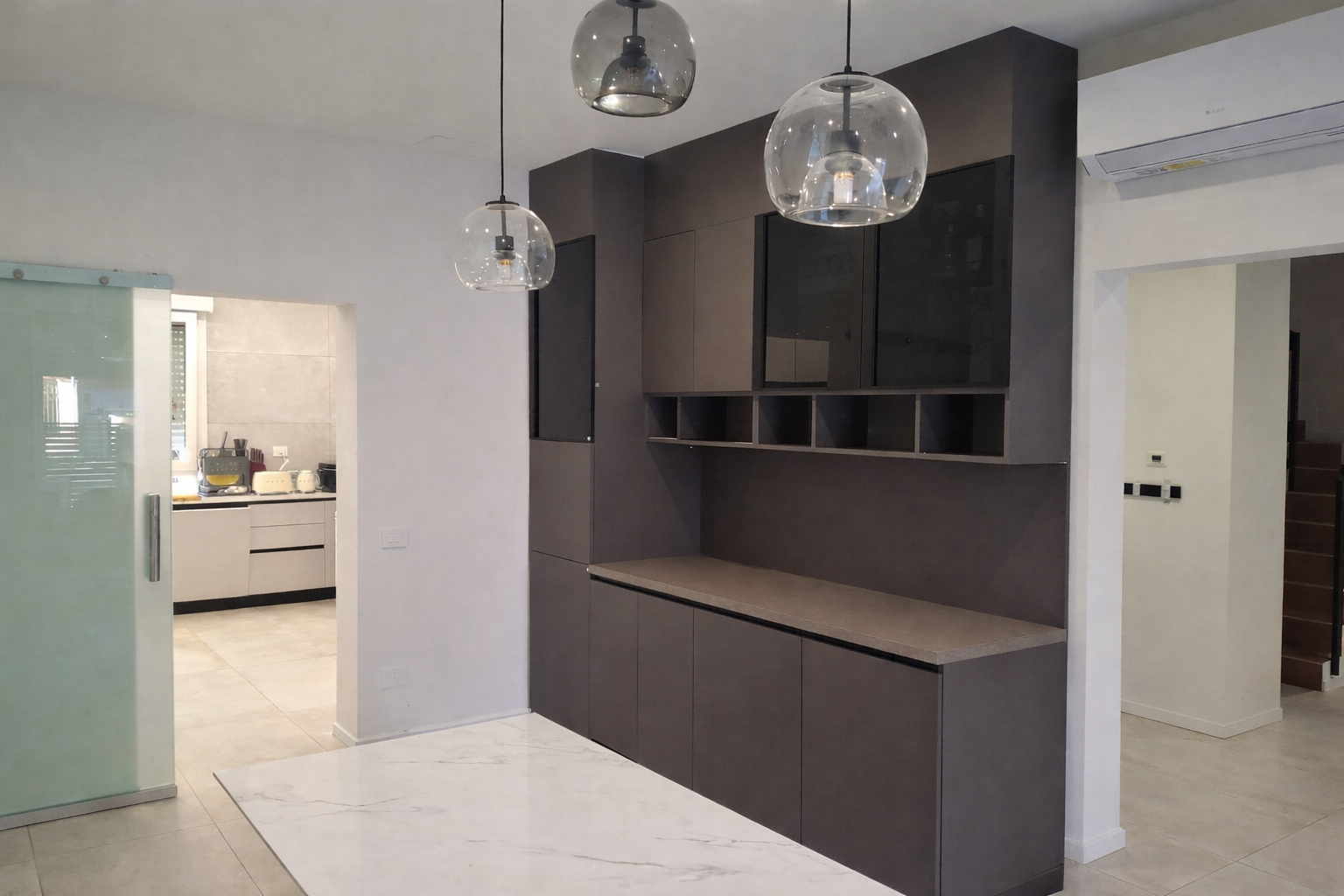 Una cucina moderna con mobili scuri, lampade a sospensione a globo e aria condizionata. Sullo sfondo si vede una scala.