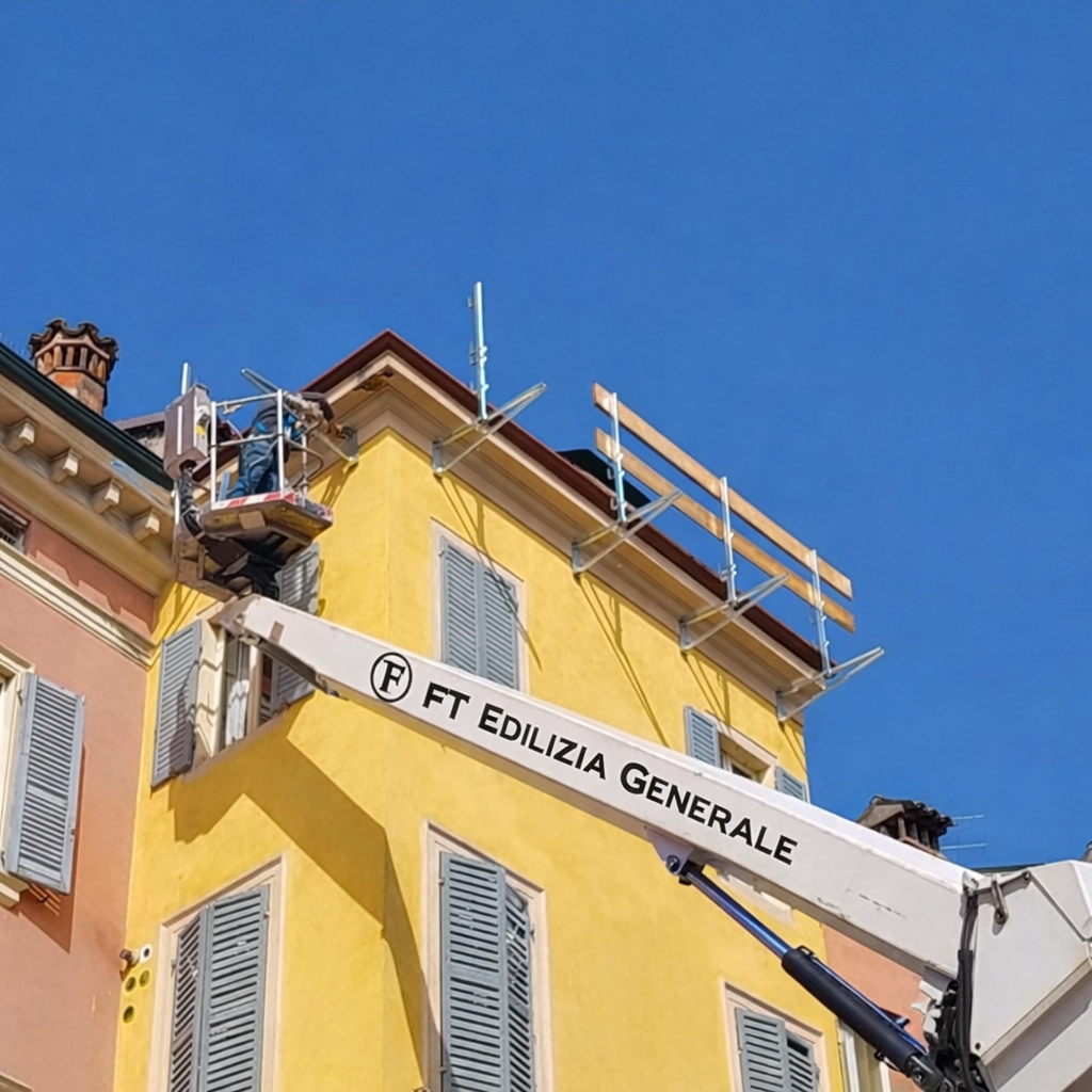 Un operaio in ascensore ripara il tetto di un edificio giallo con persiane blu, sotto un cielo azzurro e terso.