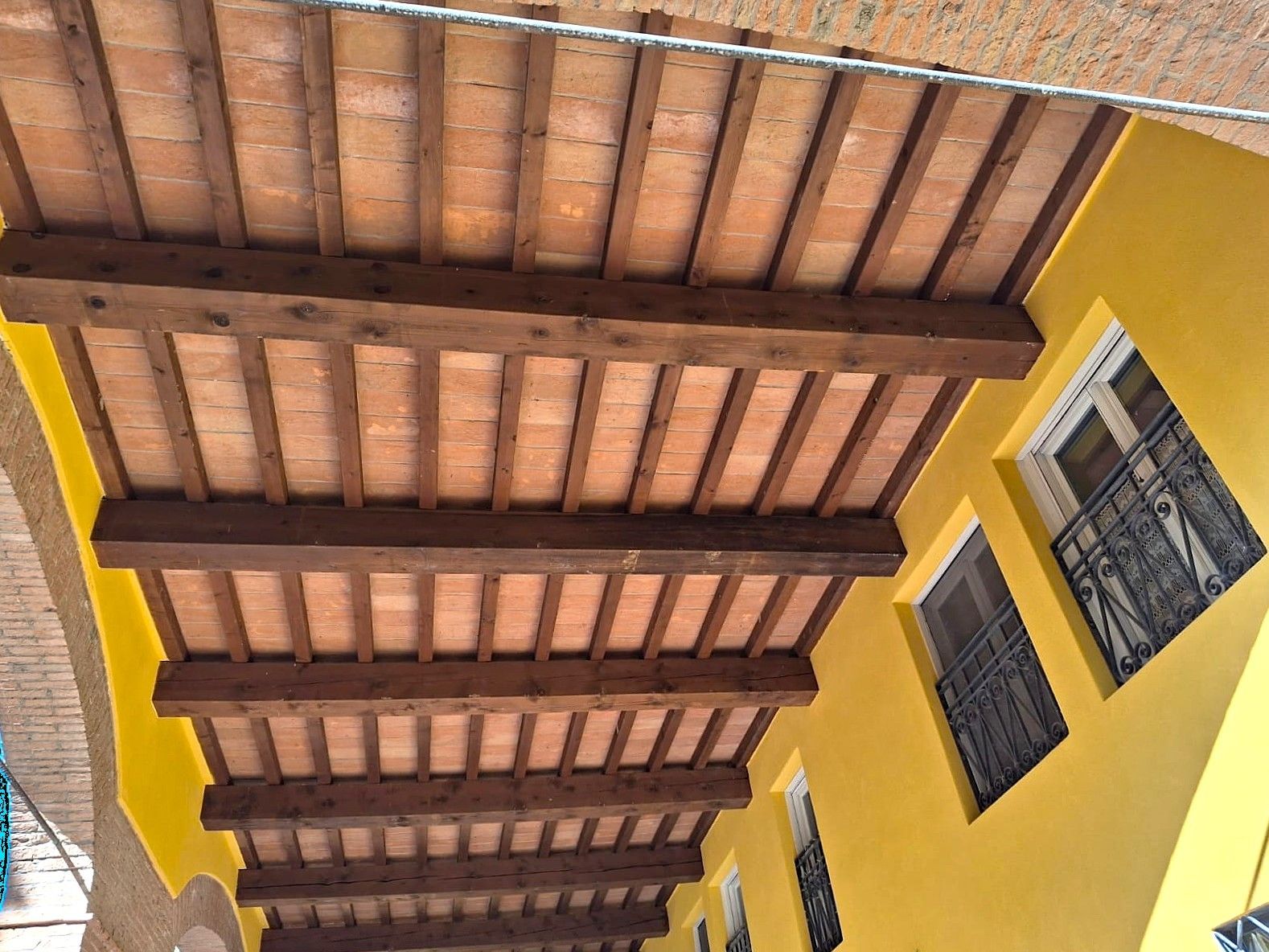 Travi in legno e tegole in terracotta formano il soffitto di un edificio dalle pareti gialle, con finestre visibili.