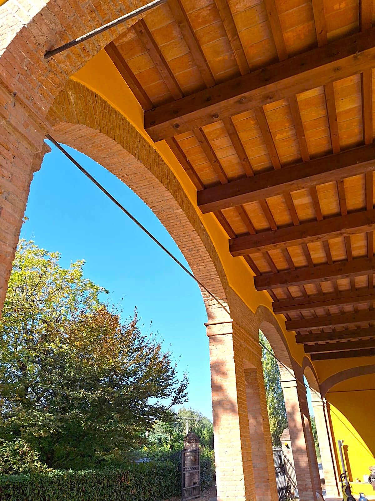 Archi e travi in mattoni sotto un soffitto giallo, che incorniciano un cielo azzurro e un albero.