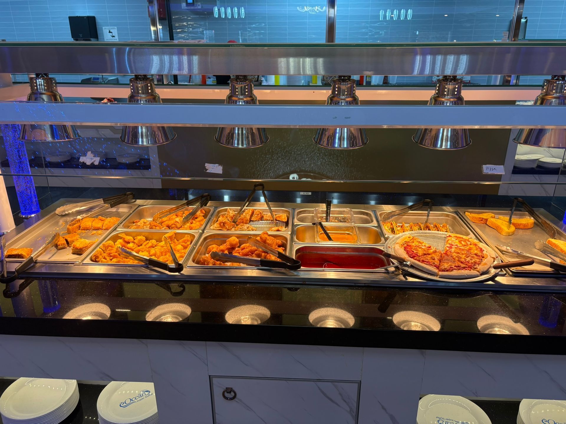 Buffet food station with trays of hot dishes under a glass sneeze guard, including noodles and sauces