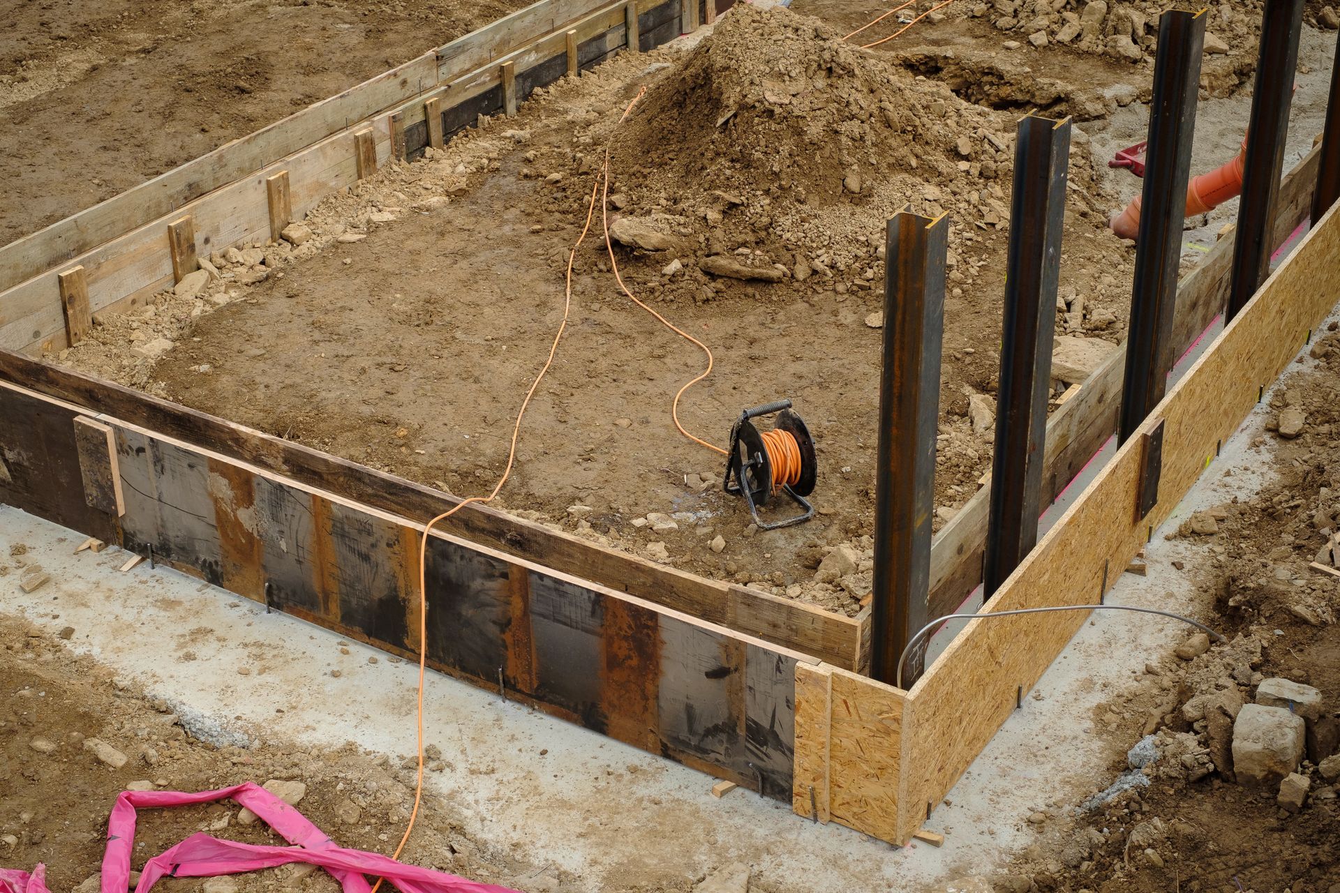 Coffrages en bois posés pour une fondation en béton sur un chantier de construction, avec remblai de terre et quelques câbles électriques présents.