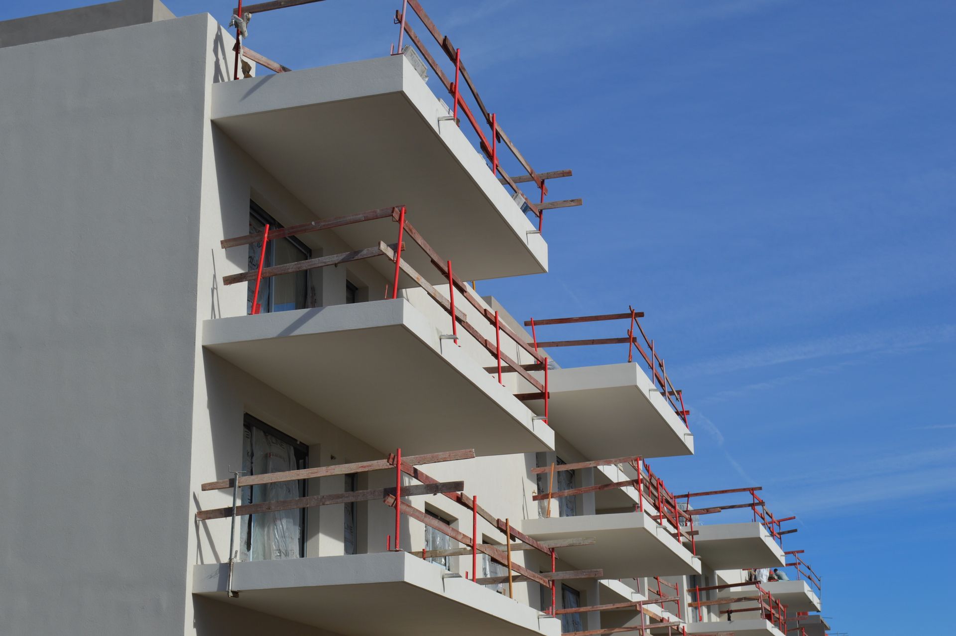 Immeuble d'appartements en construction ; balcons en béton avec garde-corps rouges contre un ciel bleu.
