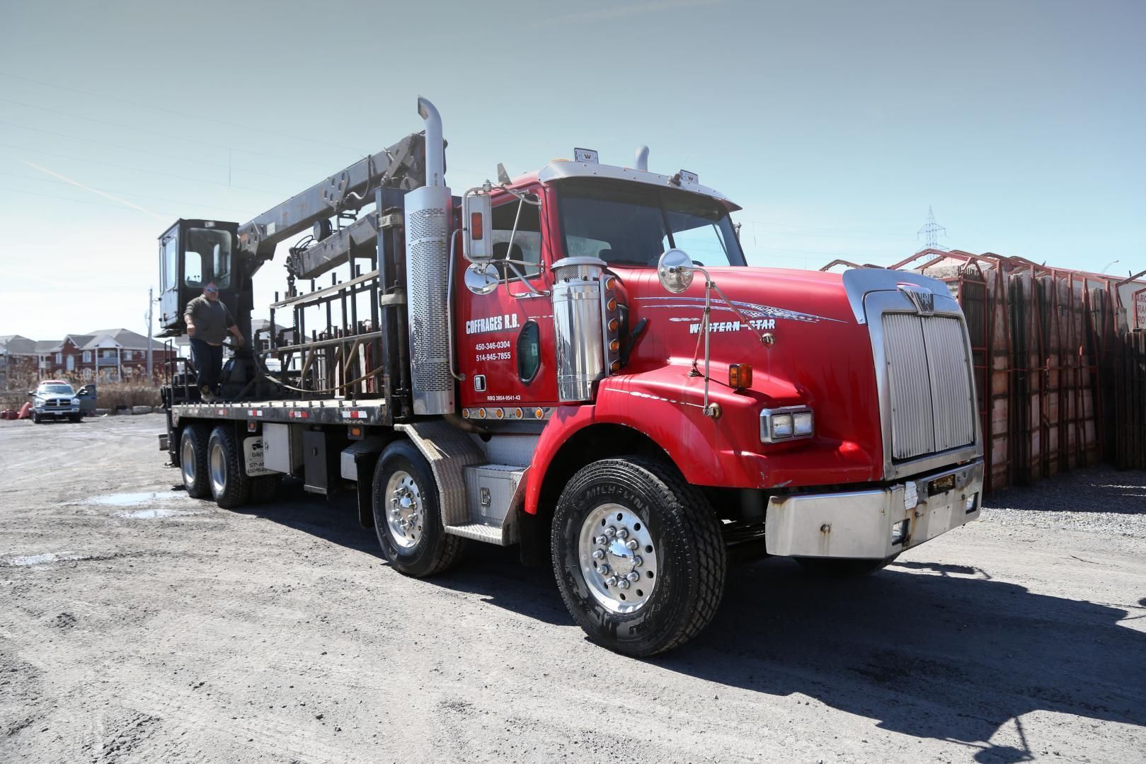 Camion plateau rouge avec grue, garé dehors, dans un endroit ensoleillé. Le camion est équipé de grandes roues et d'une calandre chromée.