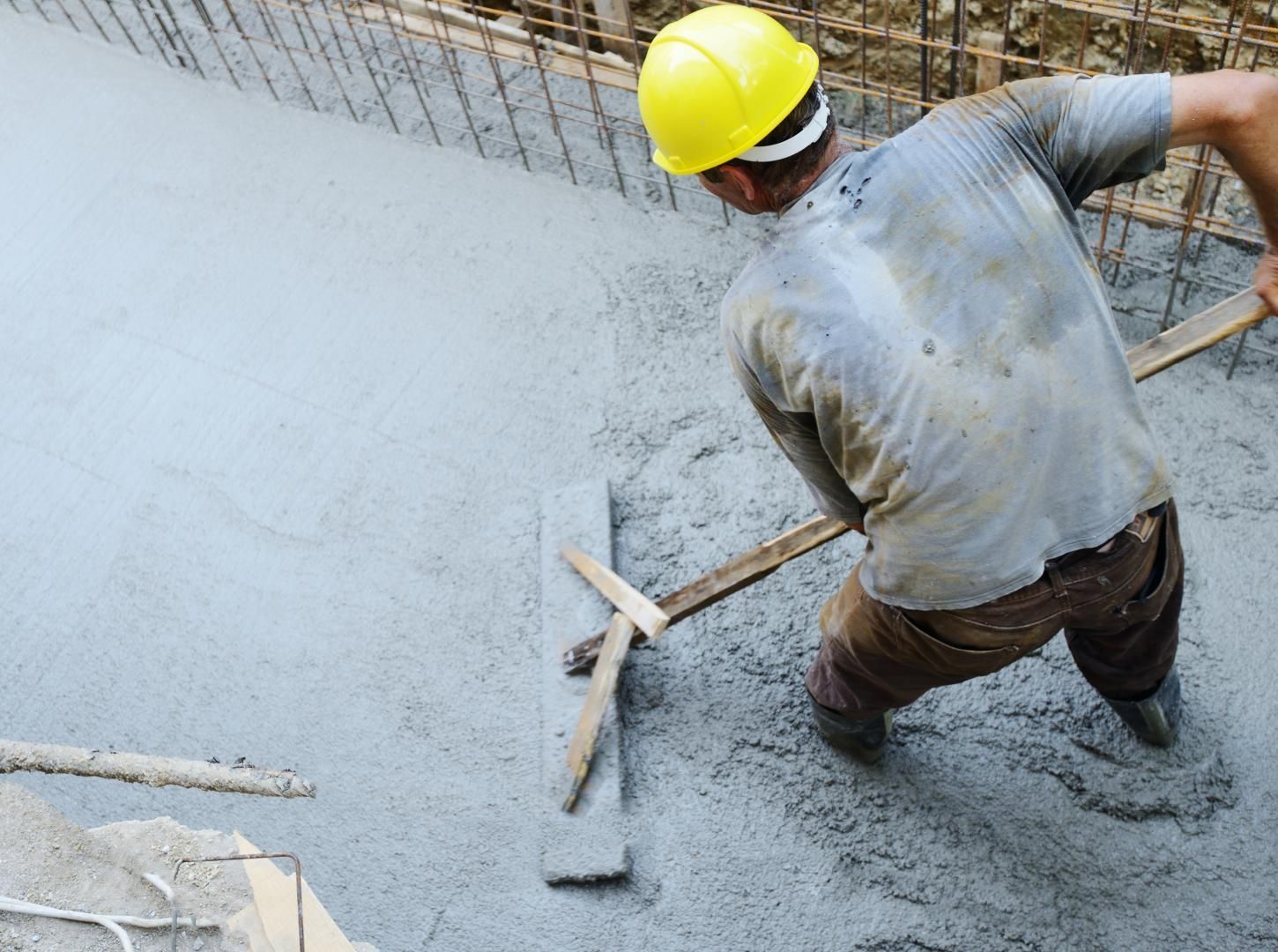 Un ouvrier du bâtiment utilise une règle pour lisser le béton humide, portant un casque de sécurité jaune.