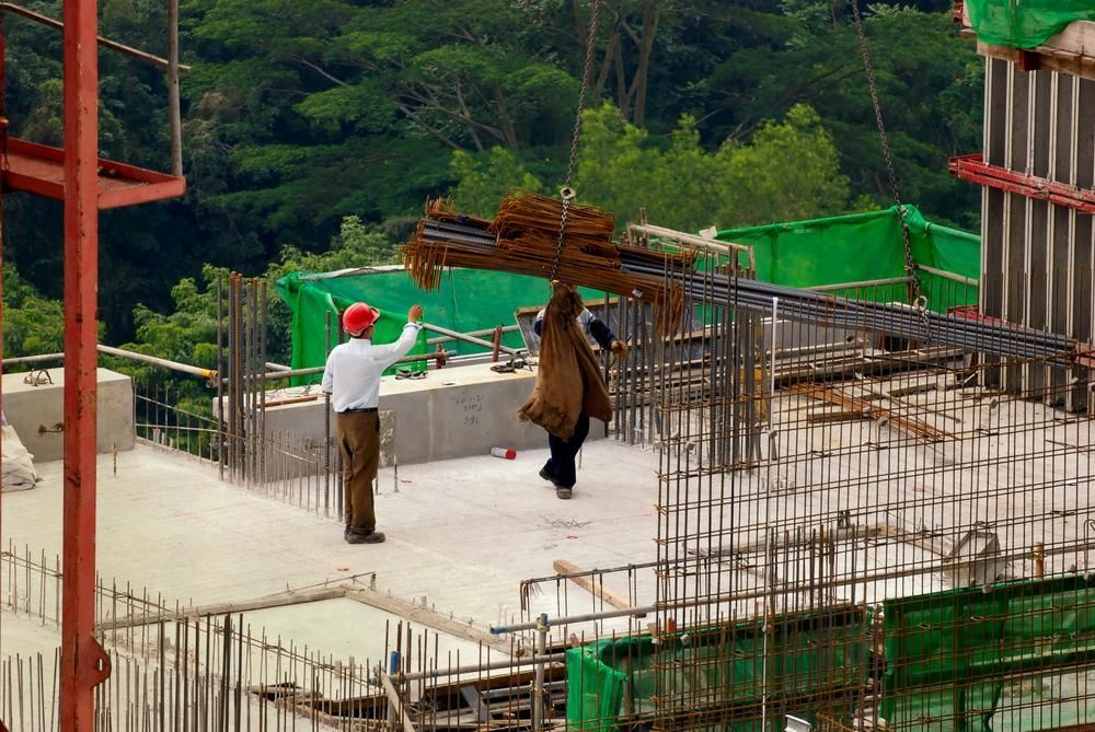 Des ouvriers du bâtiment sur un toit en béton réceptionnent des poutres d'acier d'une grue. Un ouvrier coiffé d'un chapeau rouge dirige la grue. Des arbres verdoyants en arrière-plan.