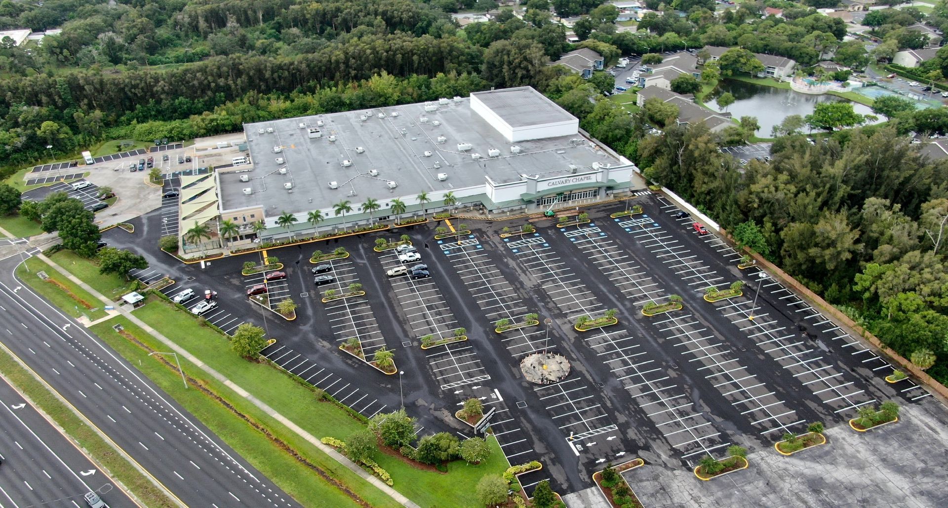 Aerial view of a large parking lot beside a warehouse, filled with many parked cars and surrounded by greenery