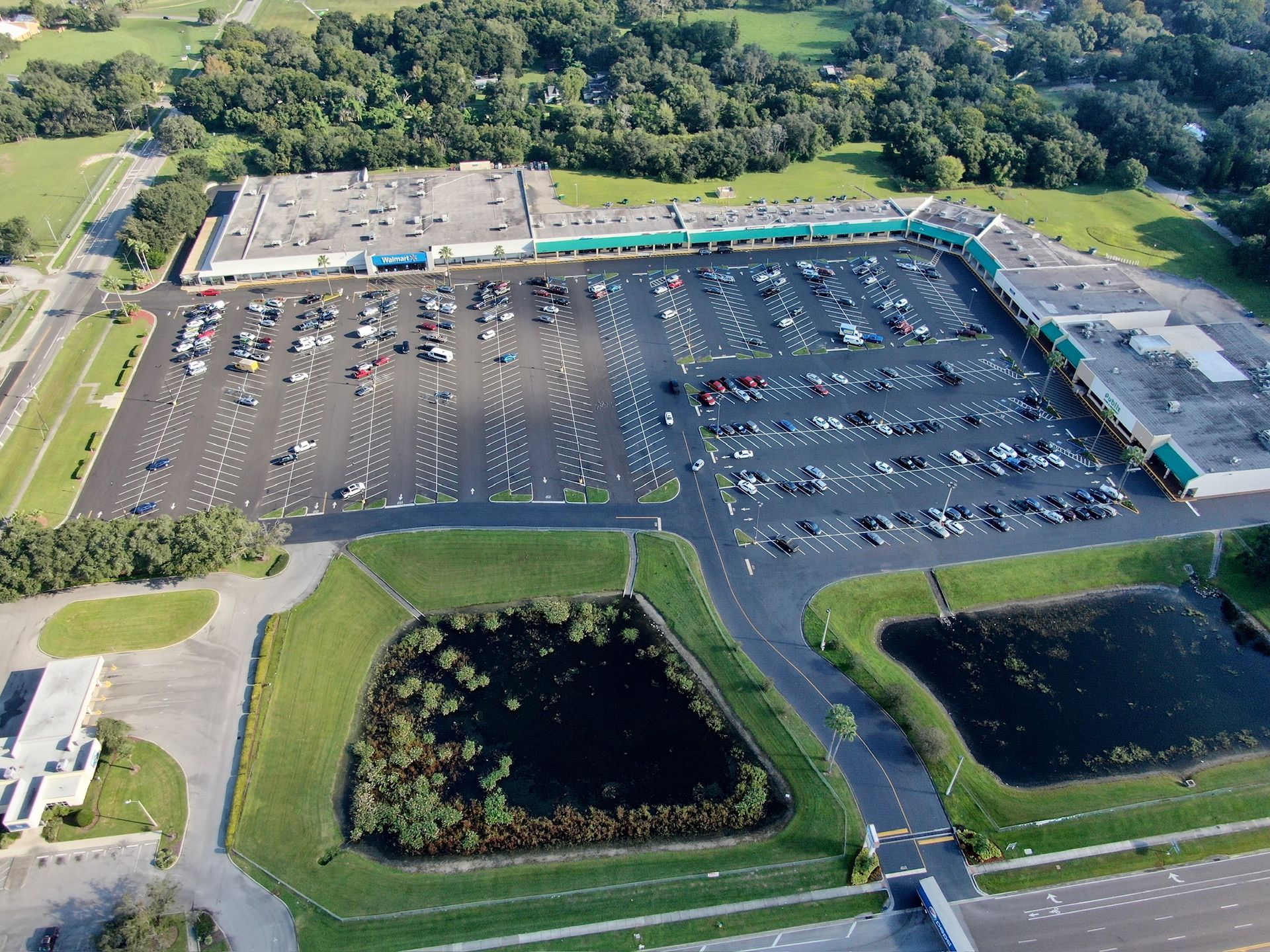 Aerial view of a large parking lot beside a commercial building, with cars and trees surrounding it.