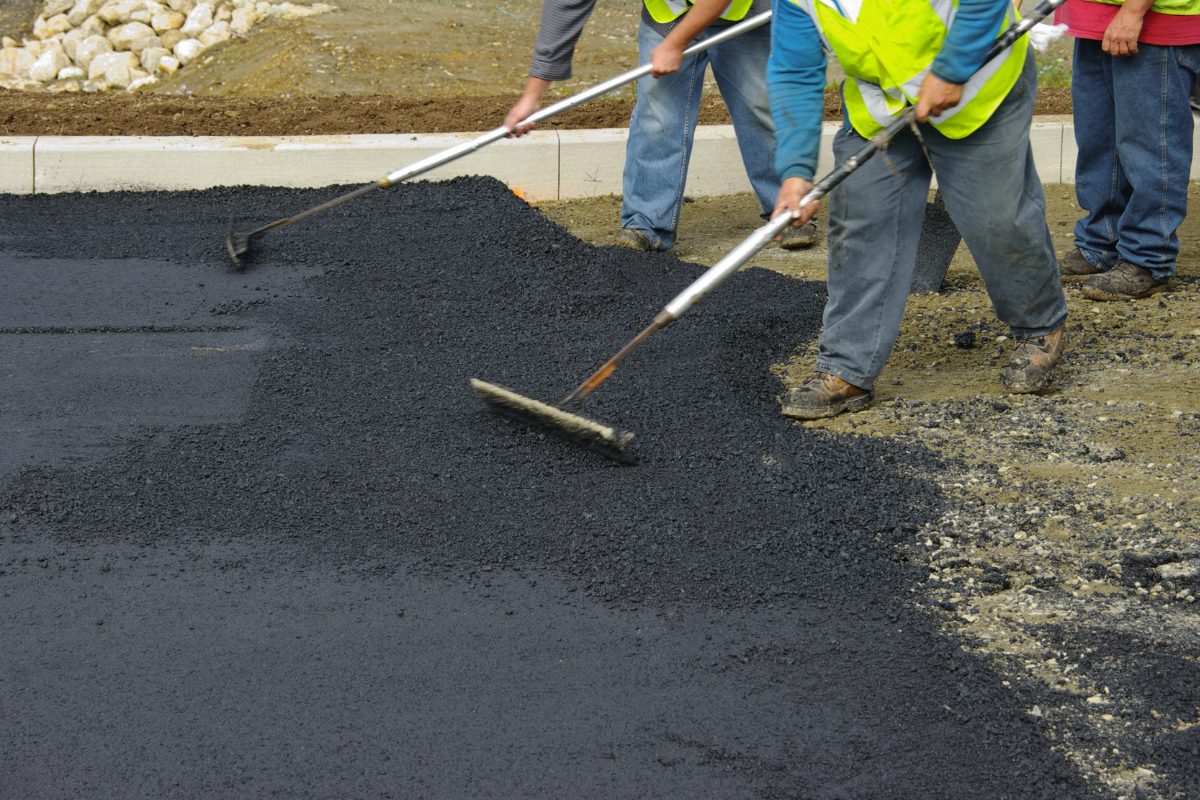Workers leveling fresh asphalt with long-handled tools on a paved surface.