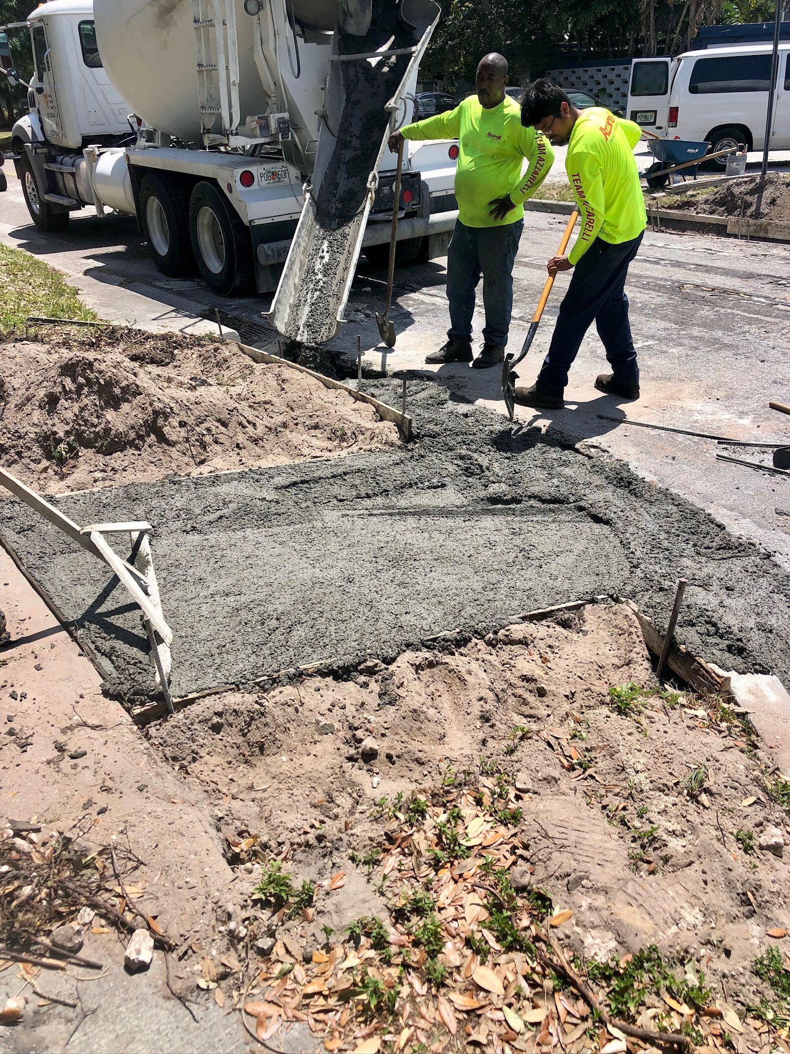 Workers in neon shirts pour concrete beside a cement truck on a road repair site.