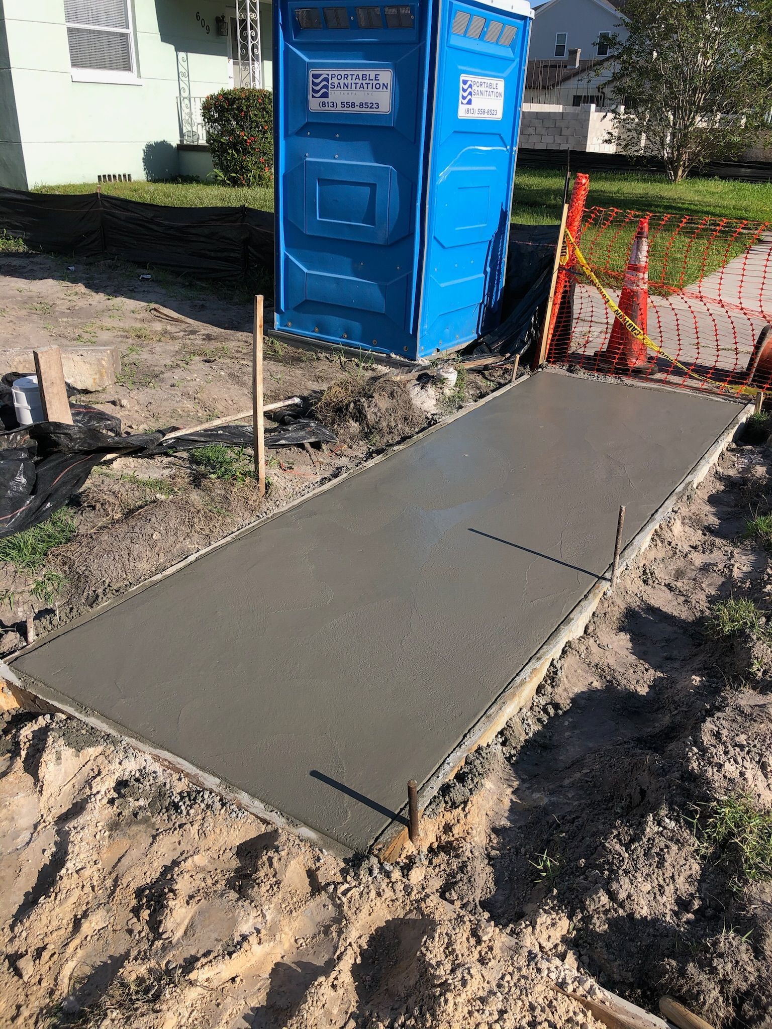 Fresh concrete sidewalk ramp beside a blue portable toilet and orange safety fencing at a worksite.