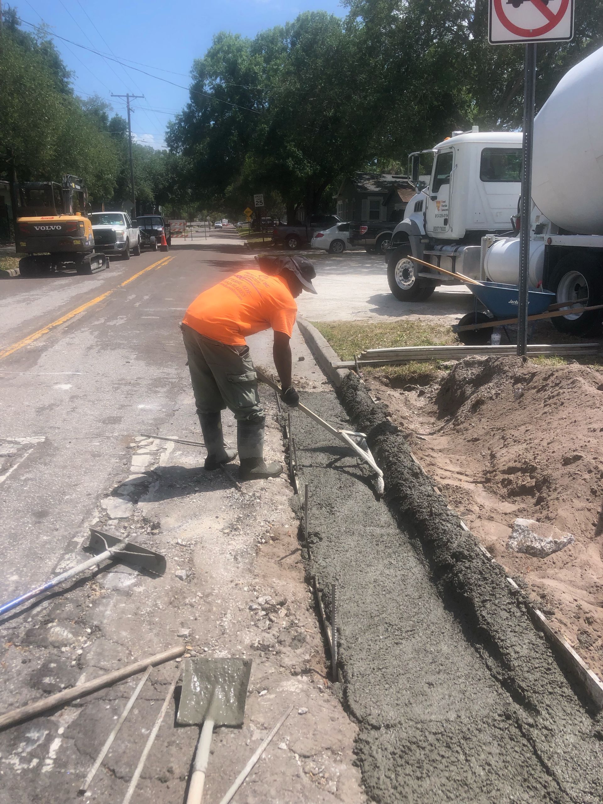 Worker in orange shirt pouring concrete into a roadside trench beside a parked cement truck.