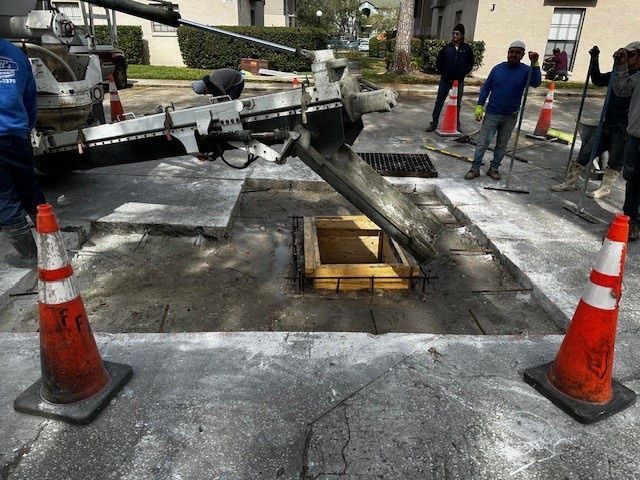 Roadwork scene with a truck lifting a heavy beam over a manhole, workers nearby and traffic cones