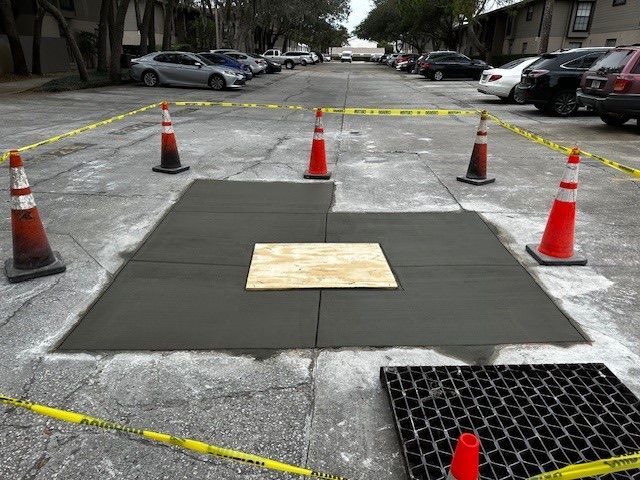 Caution-taped sidewalk repair area with traffic cones and a covered manhole in a parking lot