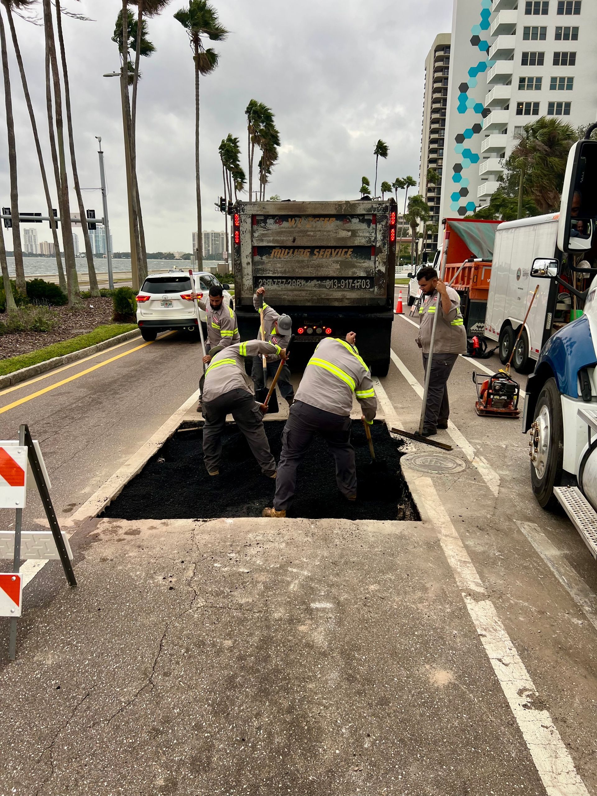 Workers repairing a pothole on a road beside a dump truck, with traffic cones and palm trees nearby