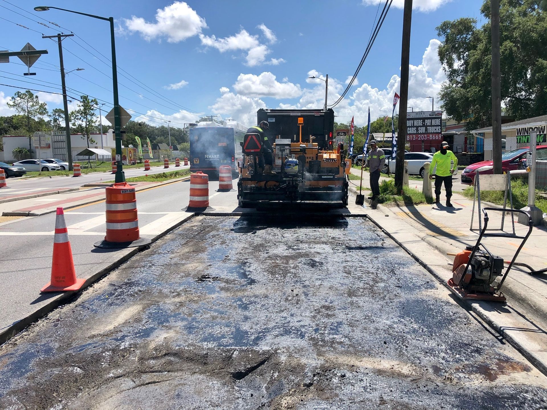 Road paving crew resurfacing a street with an asphalt paver, traffic cones, and workers in safety vests.