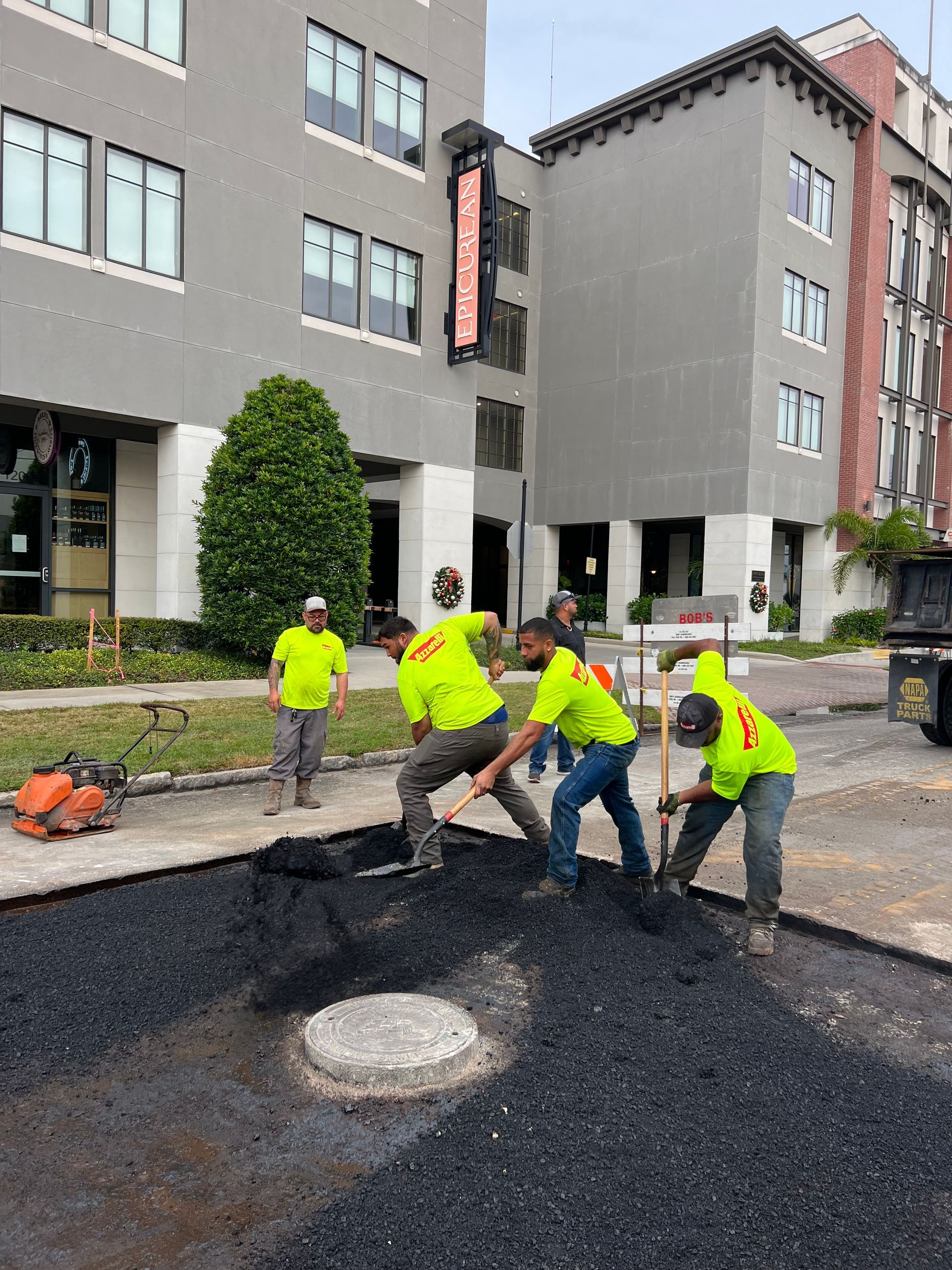 Workers in neon safety gear paving asphalt outside a gray apartment building.