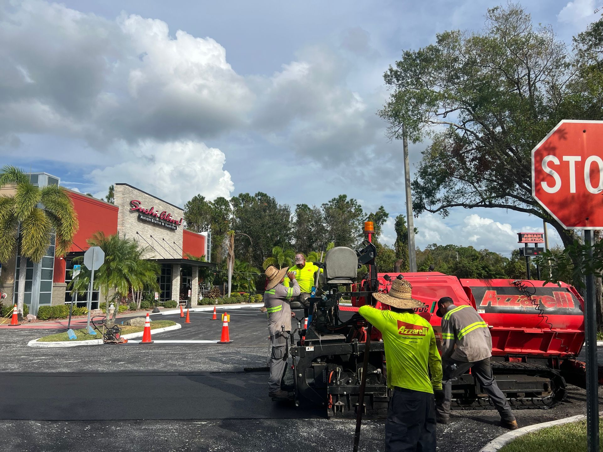 Road work crew paving a street near a stop sign, with cones, machinery, and a truck loaded with red material.