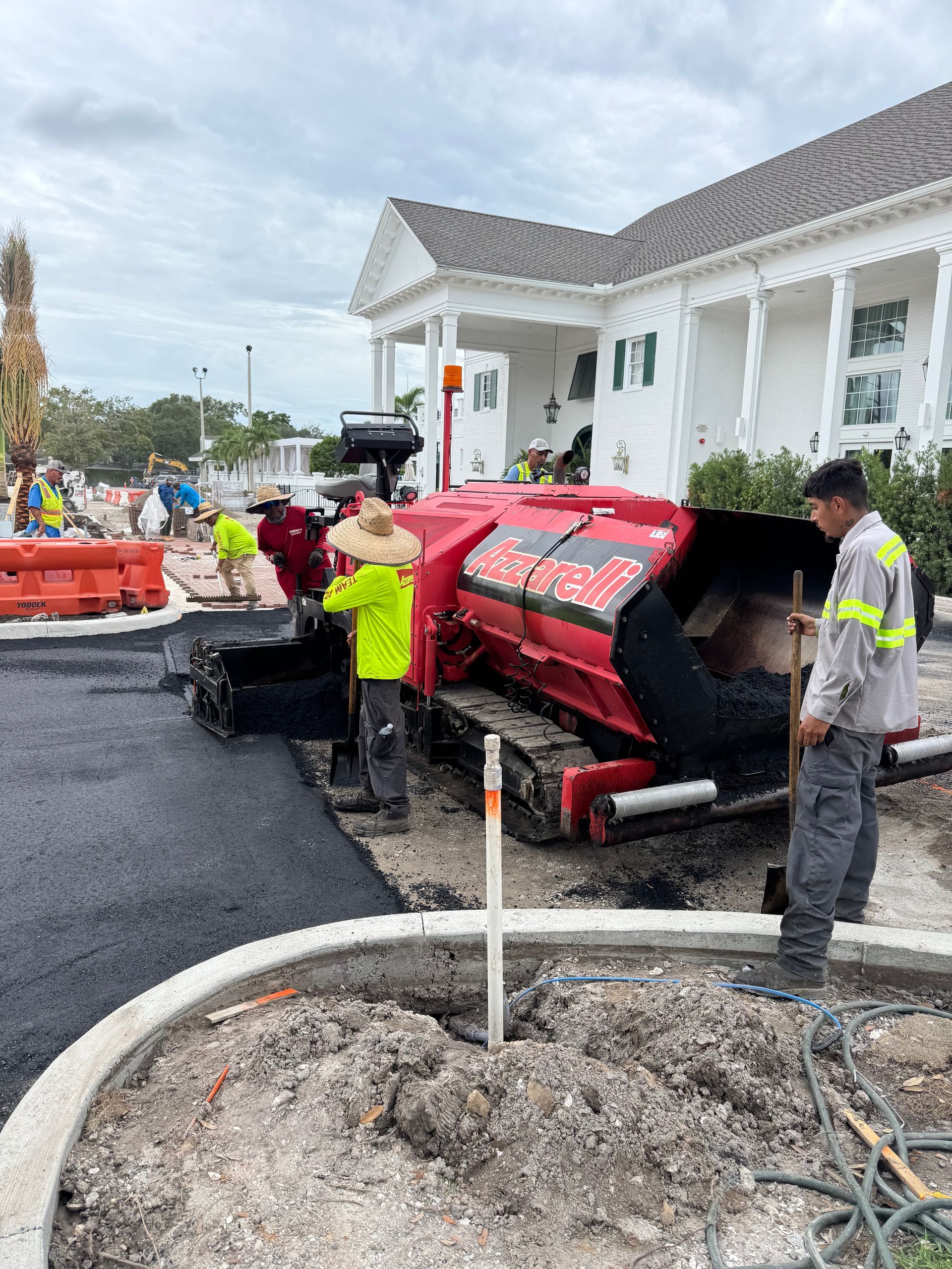 Road paving machine resurfacing a driveway beside a white house, with workers nearby.