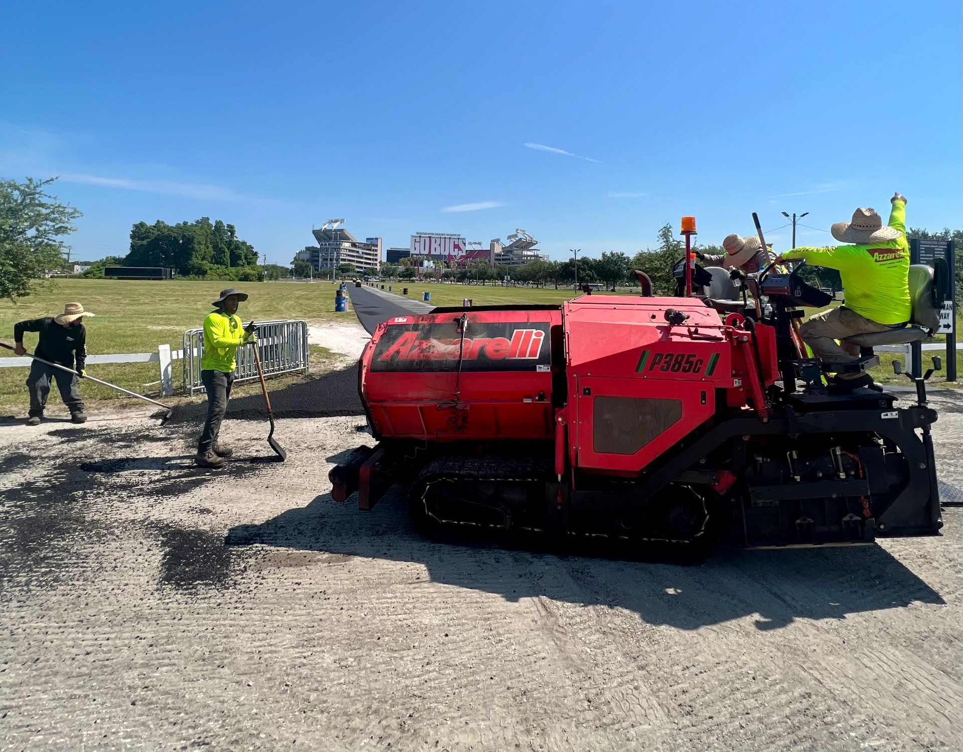 Red asphalt paver on a road site with workers in safety vests under a clear blue sky