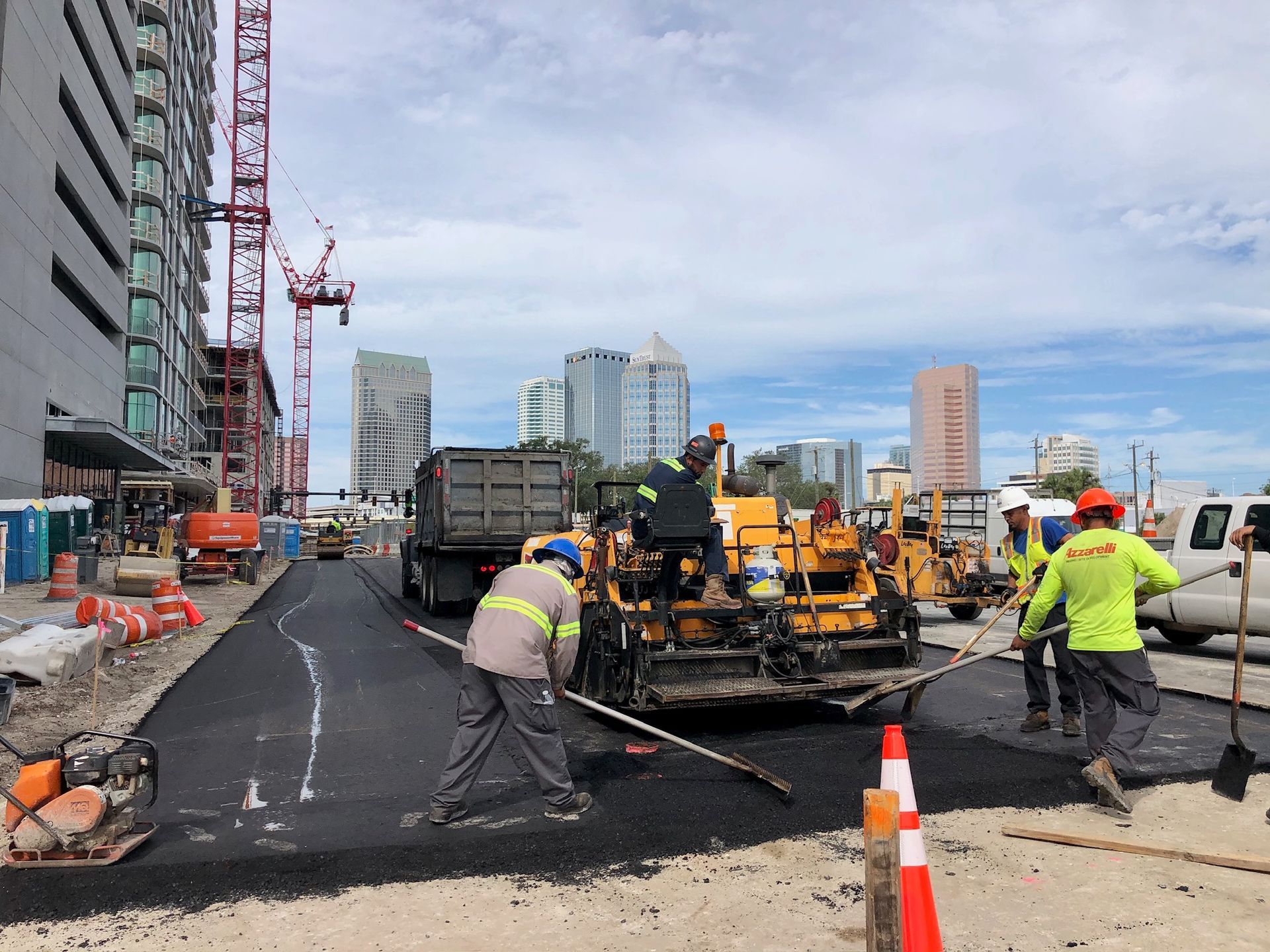Road workers paving a city street beside buildings and traffic cones, with a paving machine and trucks.