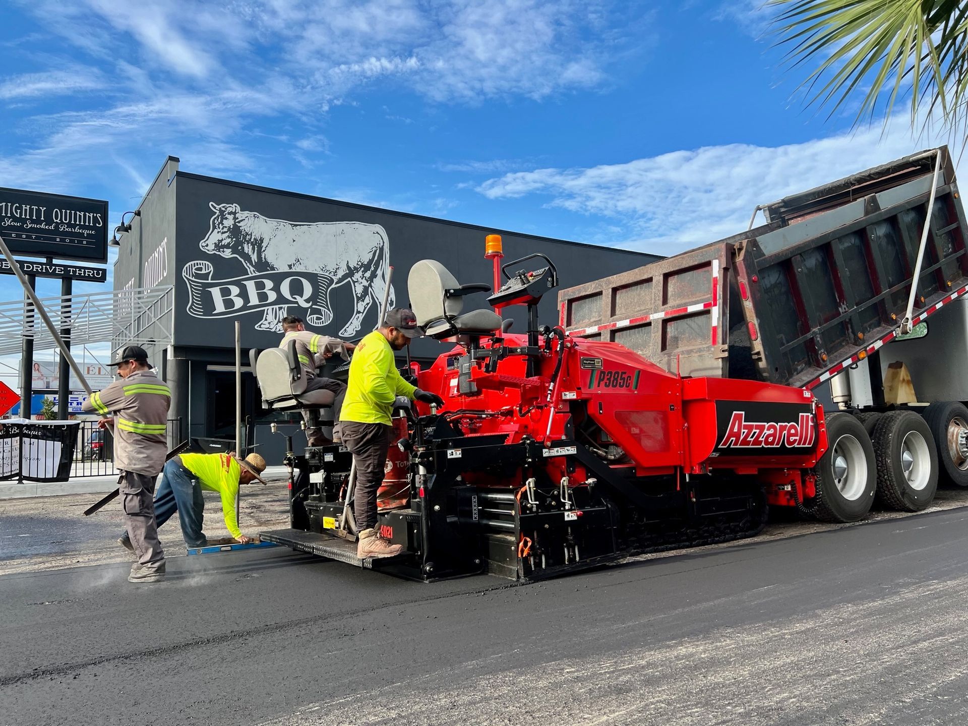 Workers operate a red asphalt paver beside a dump truck outside a BBQ restaurant.