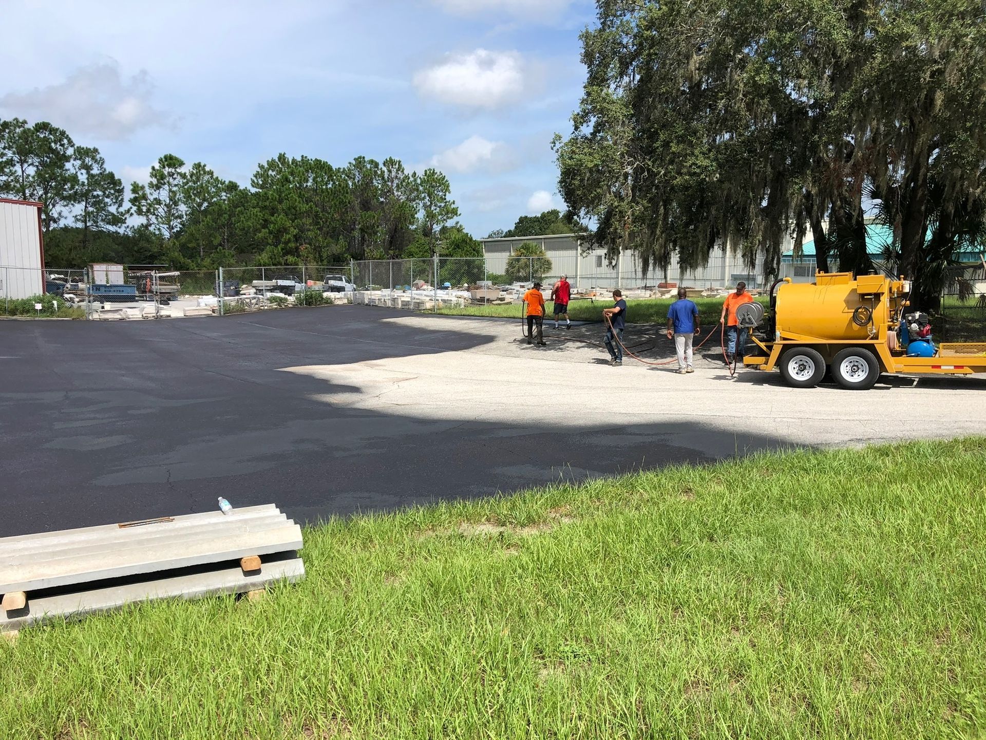 Workers paving a new blacktop area beside a grass field, with a yellow asphalt machine nearby.