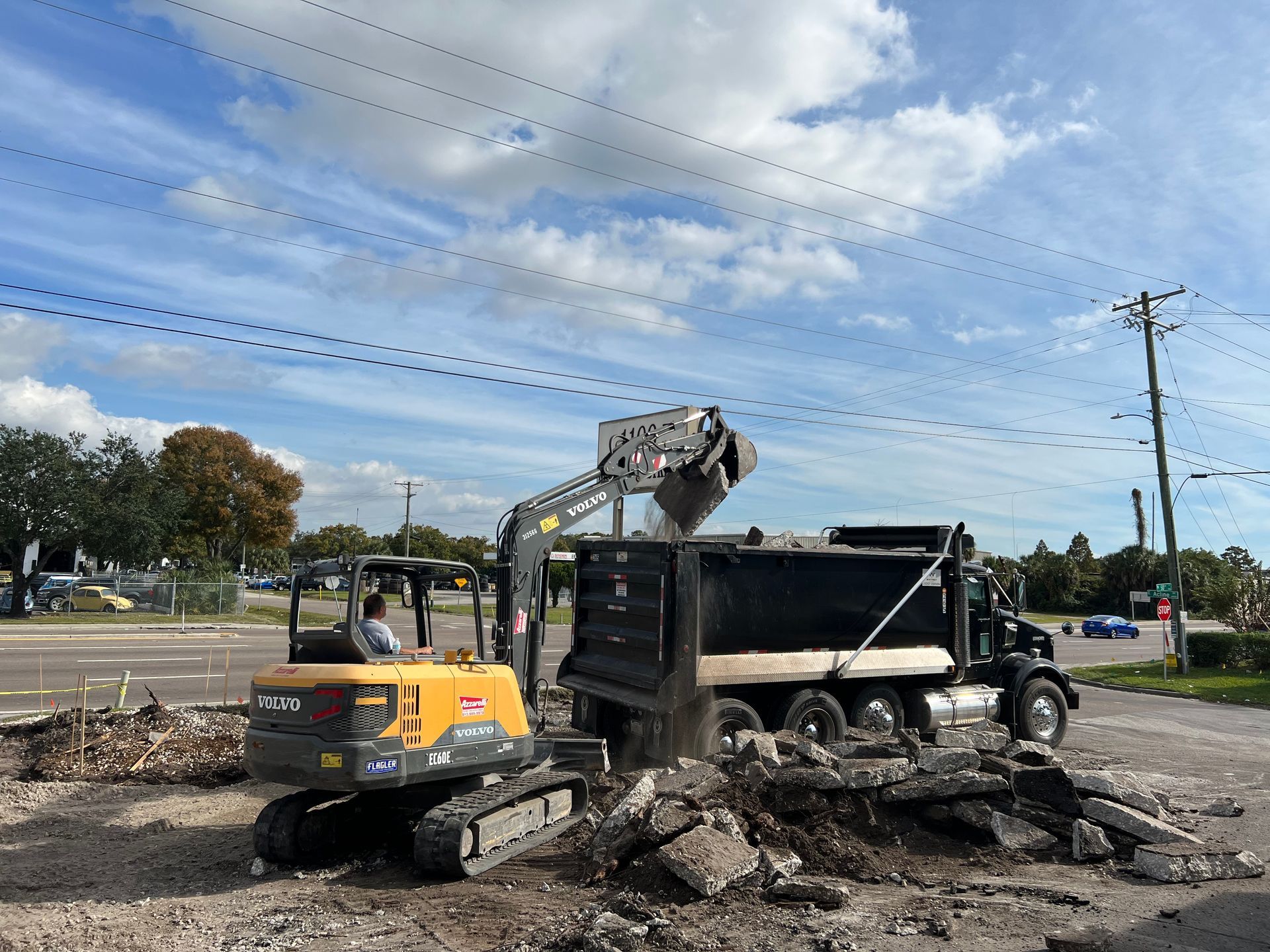 Yellow excavator loading rubble into a black dump truck at a roadwork site under a blue sky