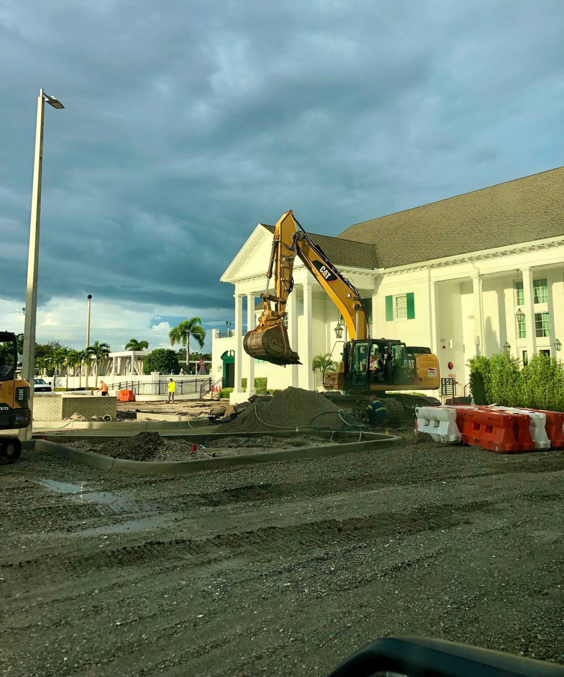 Excavator digging at a building site beside a white house under cloudy skies