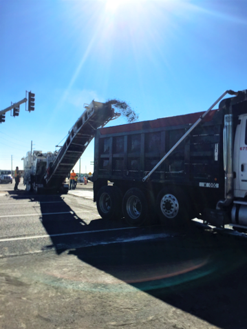 Asphalt milling machine loading debris into a dump truck on a street under a bright sun.