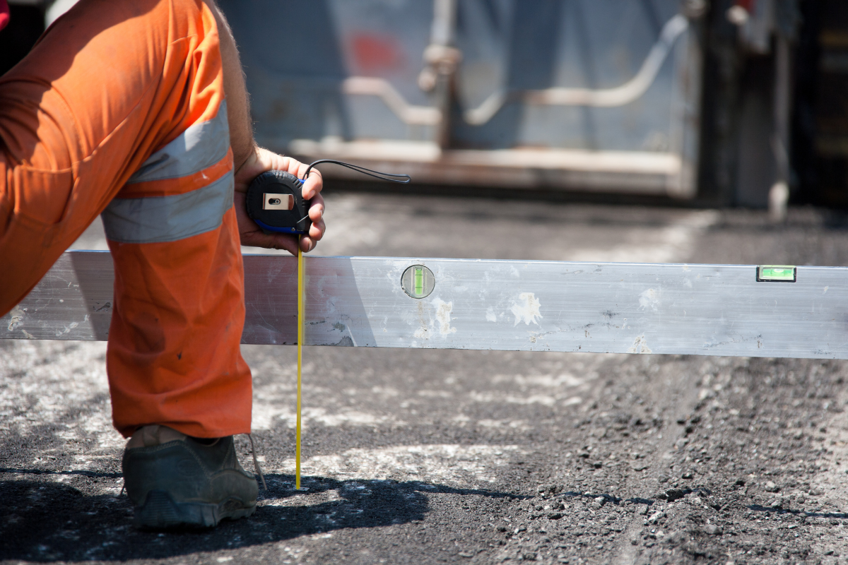 Construction worker in orange overalls measuring asphalt with a level.