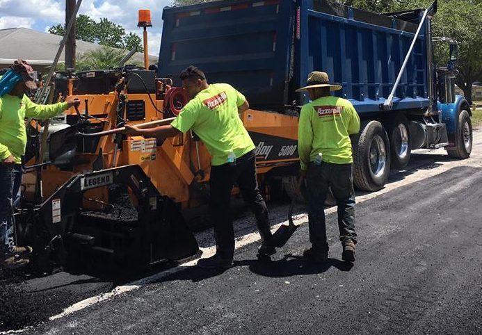 Road crew paving road; dump truck pouring asphalt into paving machine. Workers in neon green shirts.