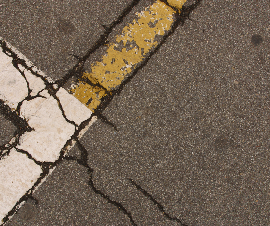 Cracked asphalt with yellow and white painted lines, likely a road or parking lot.