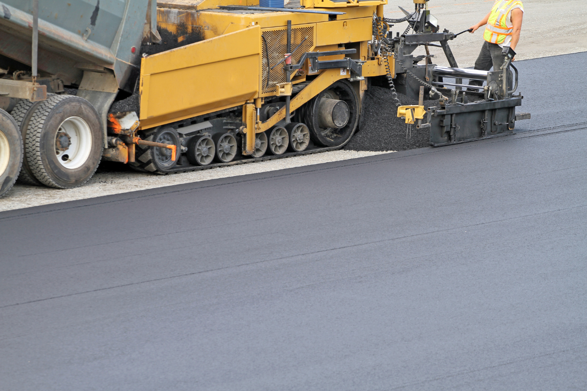 Asphalt paving machine laying fresh black pavement; worker in safety vest.