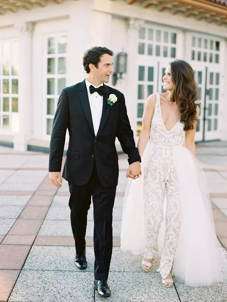 A bride and groom are walking down a sidewalk holding hands.