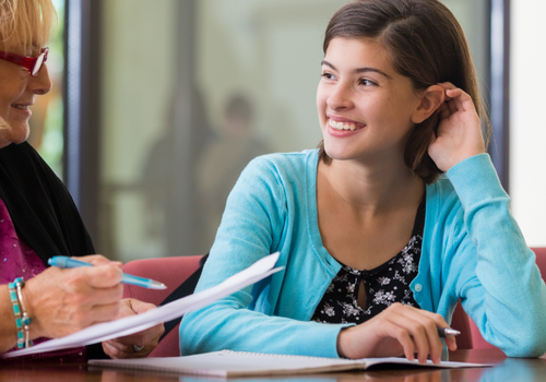 Two women in a classroom talking over papers, one smiling with a pen and laptop
