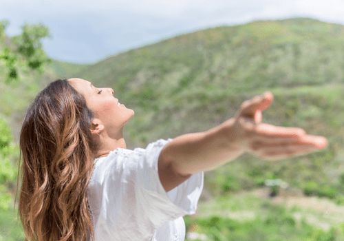 Woman in white shirt with arms outstretched, smiling outdoors in a green hilly landscape