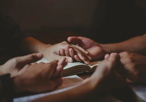 Hands gathered around a book in a dim, warm-lit setting, reading or studying together