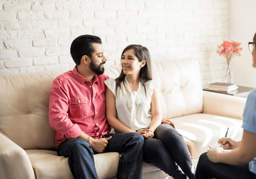 Two people talking on a beige couch in a bright living room.