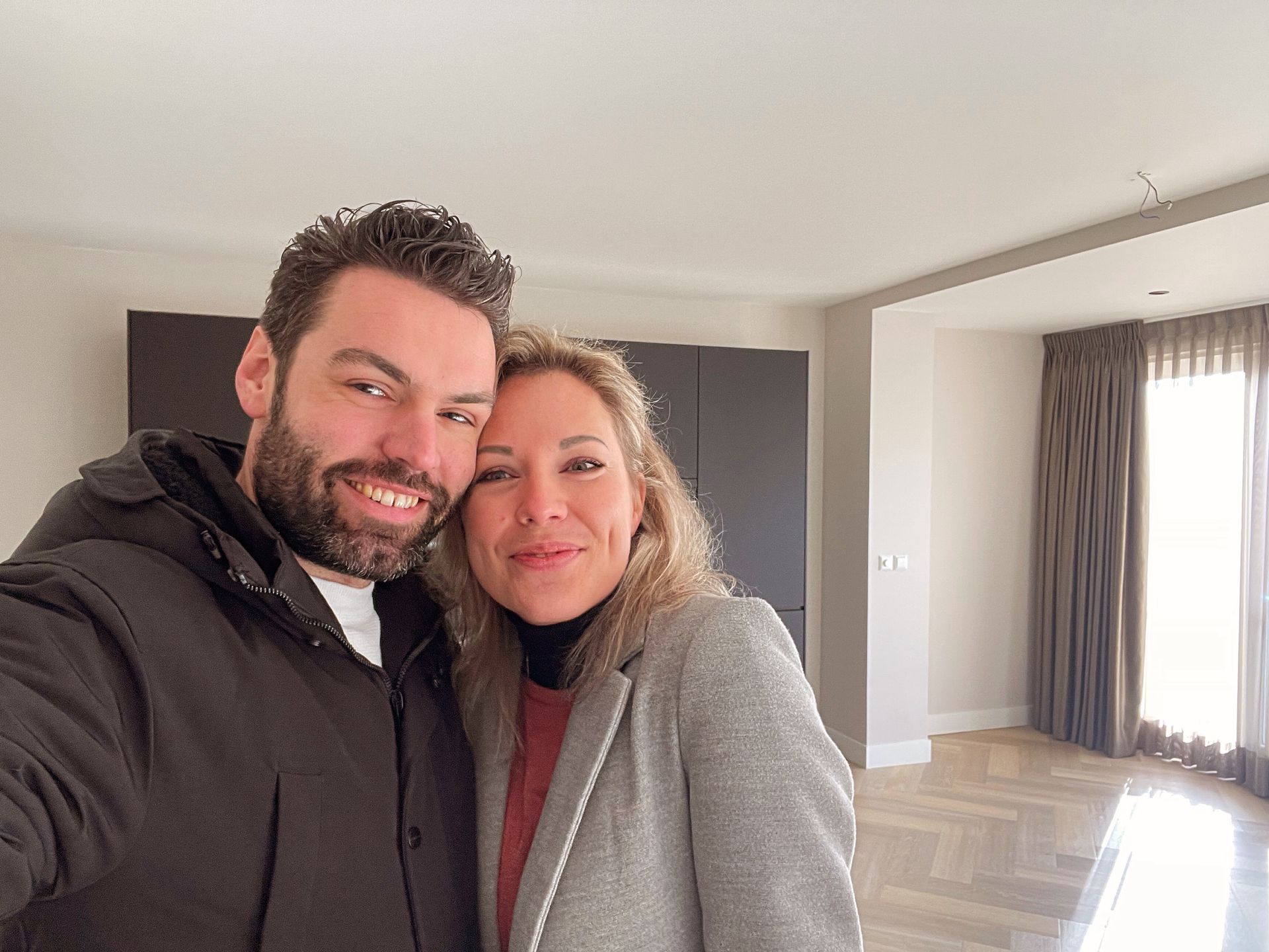Een lachend stel poseert in een lege kamer met donkere kasten en een raam met gordijnen.