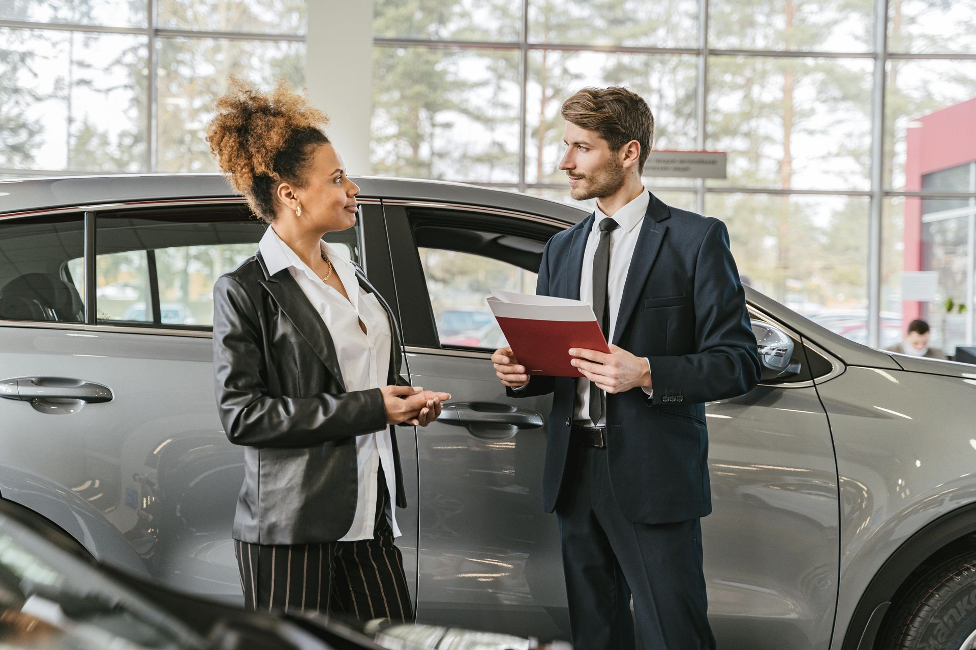 Vrouw en man bij autodealer, man houdt papieren vast, vrouw maakt gebaren, auto op de voorgrond.