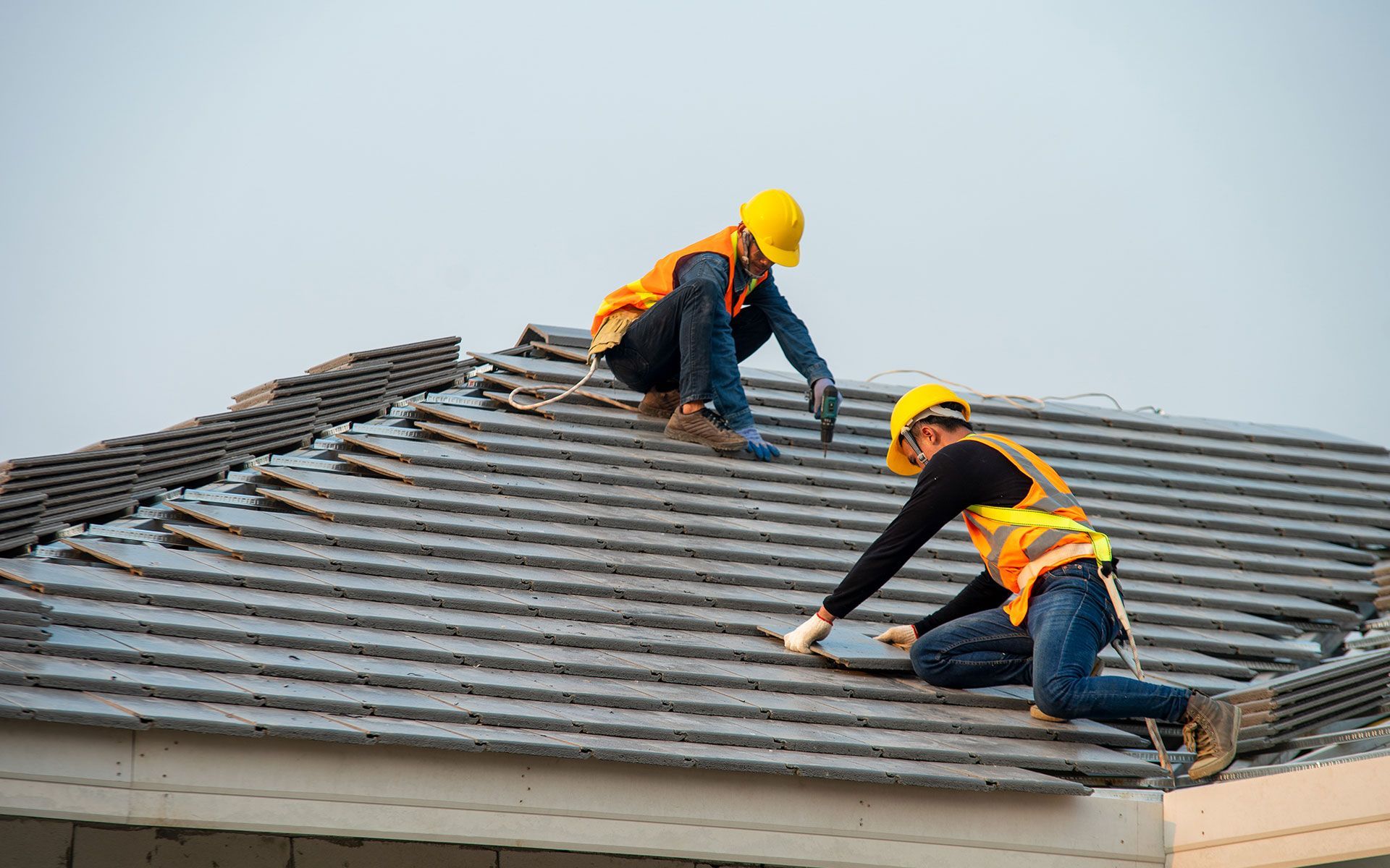 Two roofers in safety vests and hard hats installing tiles on a roof.
