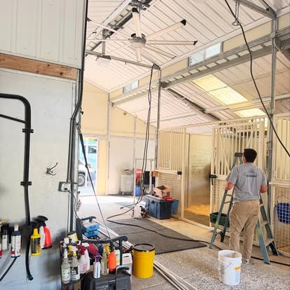 Inside a stable: man on a ladder, working near horse stalls, with fan and supplies visible.