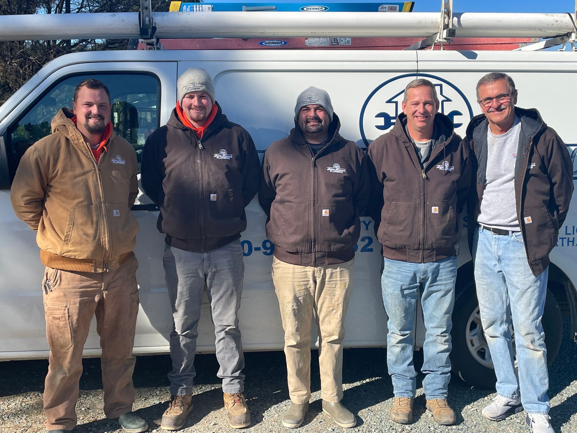 Five men stand in front of a work van, all wearing work jackets. The van has a company logo.