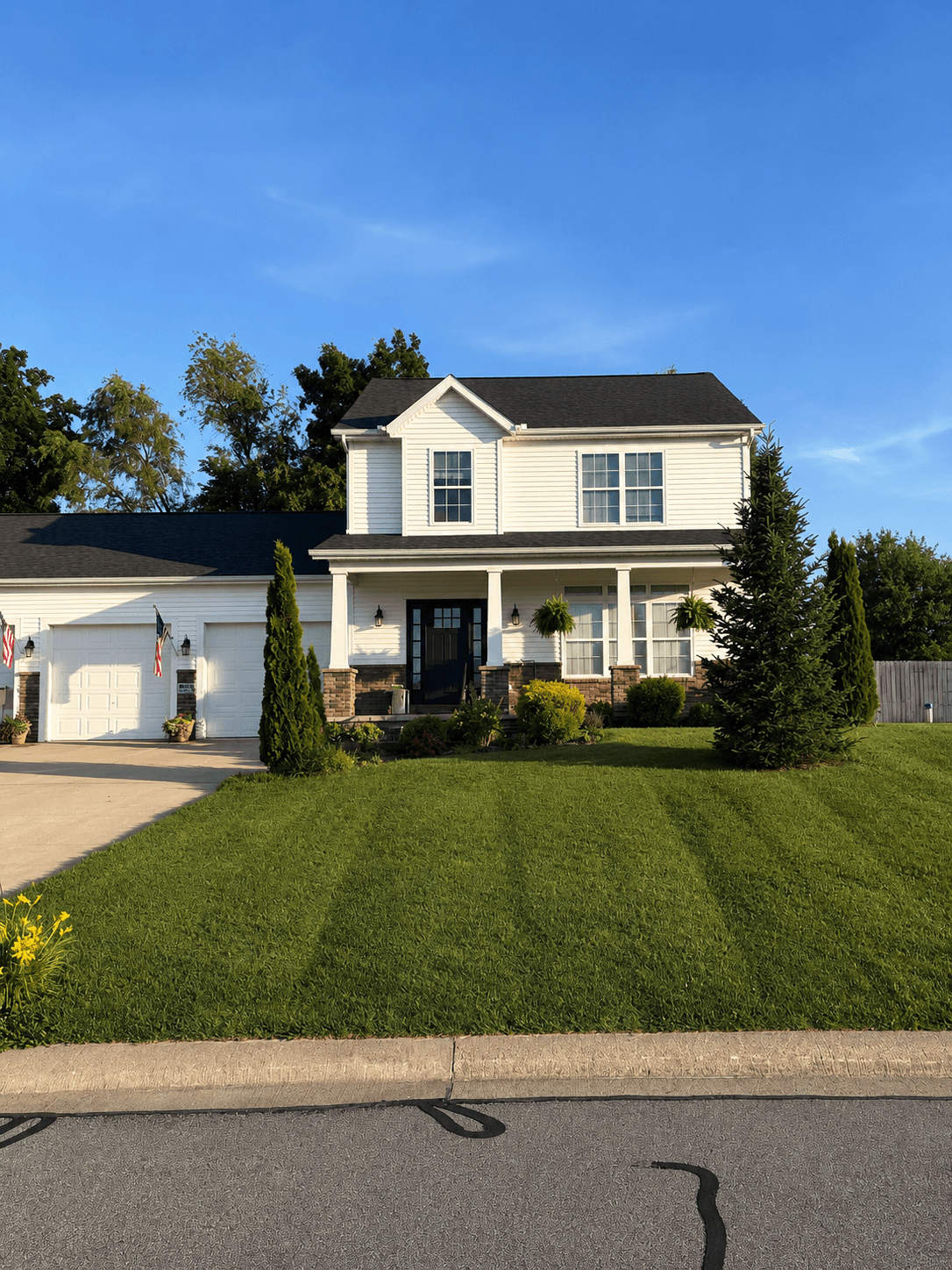 Two-story white house with black shutters, a front porch, and a green lawn on a sunny day