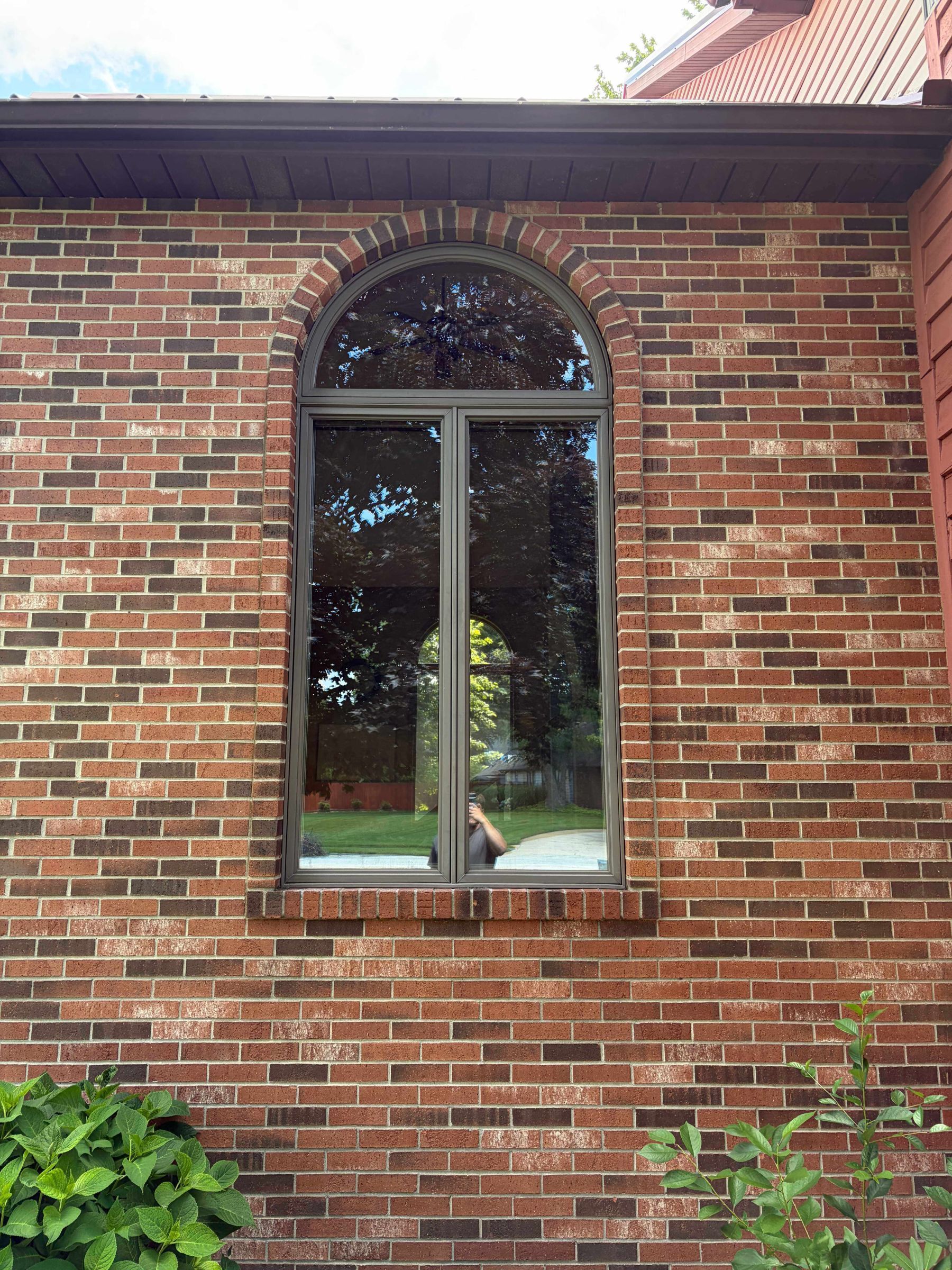 Brick house wall with arched window and red roof eave, framed by green plants.