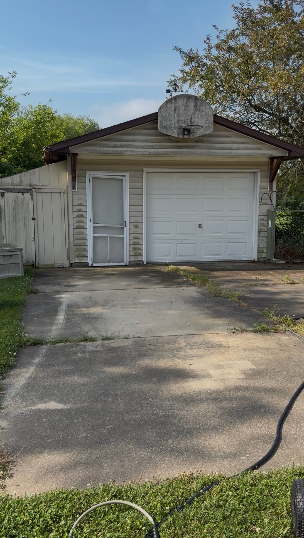 Small beige house with white garage door, driveway, and trees in the front yard