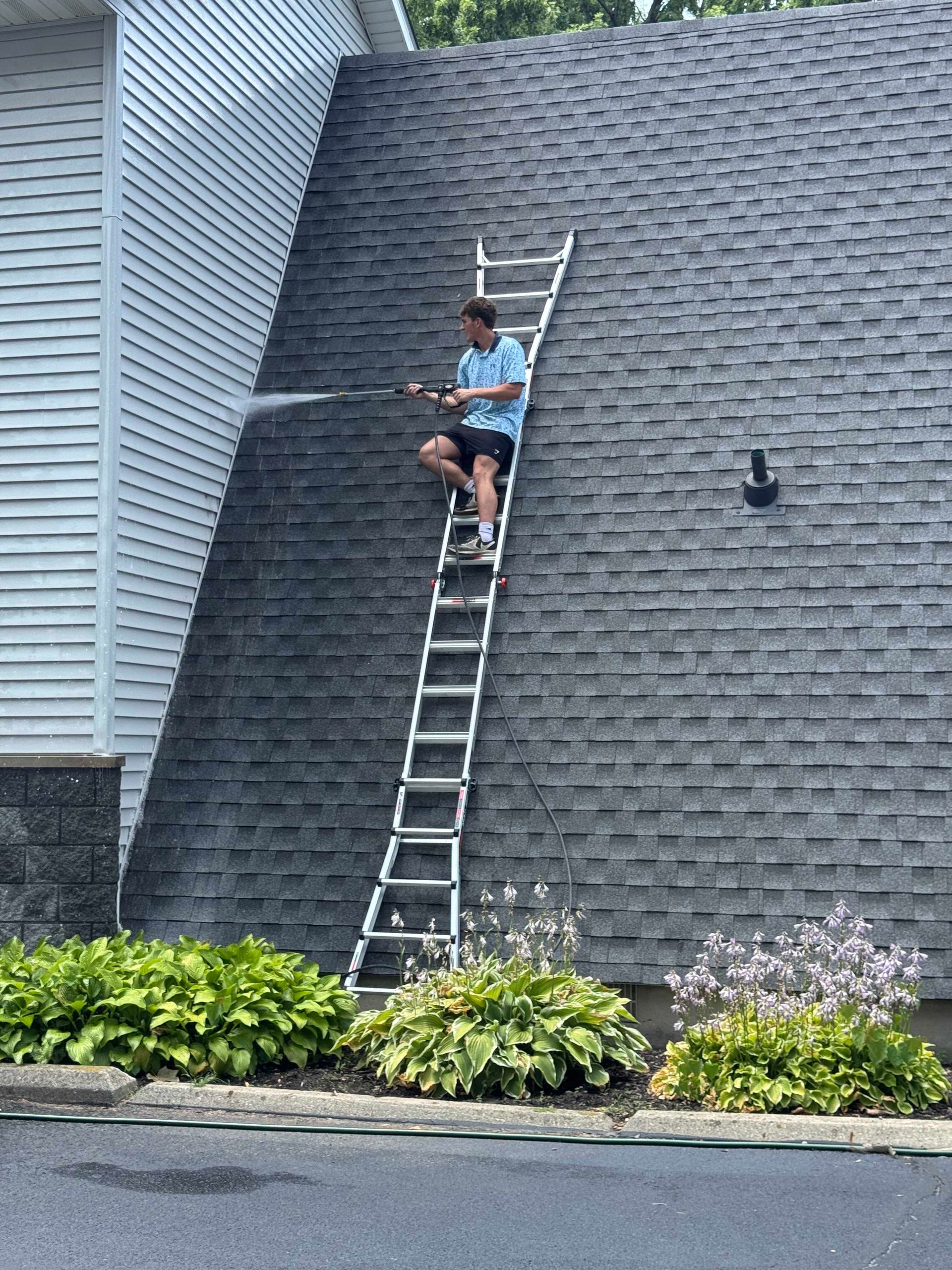 Person sitting on a ladder leaning against a gray shingled roof, adjusting something near the top.